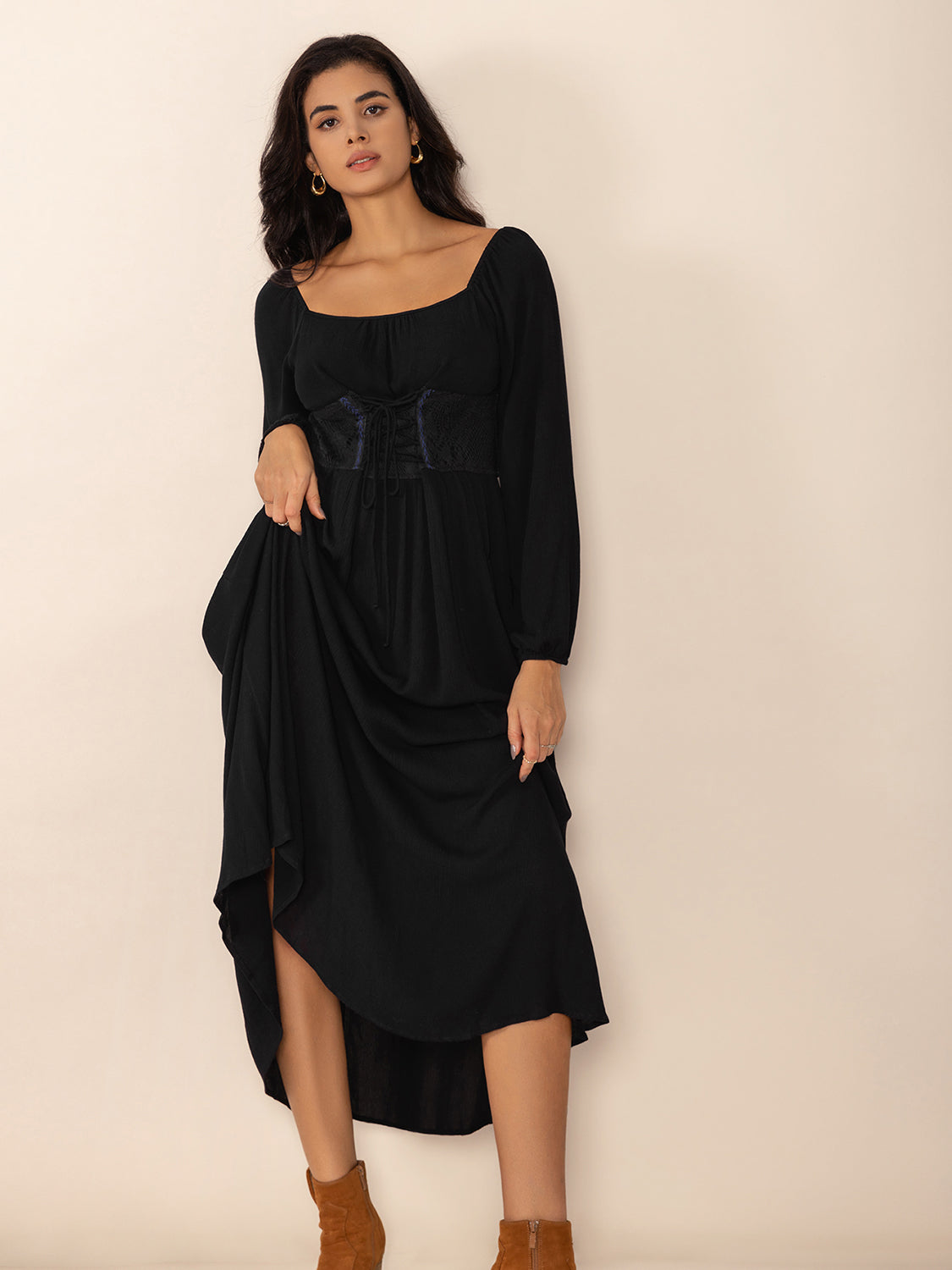 Woman wearing a black midi dress with a square neckline, long puff sleeves, and a corset-style waist detail, lifting the hem of the skirt to reveal her legs.