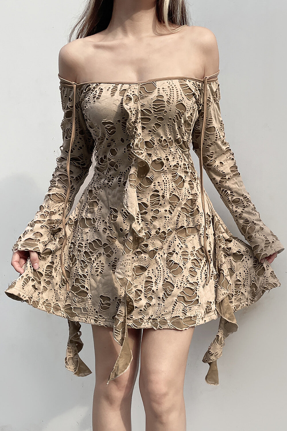 A woman stands against a white background wearing a beige distressed off-the-shoulder dress.