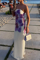 Woman wearing a white strapless maxi dress with a purple floral print, holding a silver handbag and a drink, standing on a paved walkway near a beach.