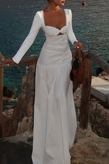 Woman wearing a white long-sleeved maxi dress with a cut-out and ruched detailing, leaning on a wooden railing by the sea.