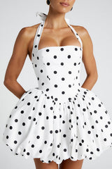 Front view of a woman wearing a white and black polka dot halter neck dress with a square bodice and a voluminous bubble hem skirt, against a white background.