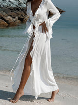Woman standing on a sandy beach with clear turquoise water, wearing a white ruffled maxi dress with a high slit.