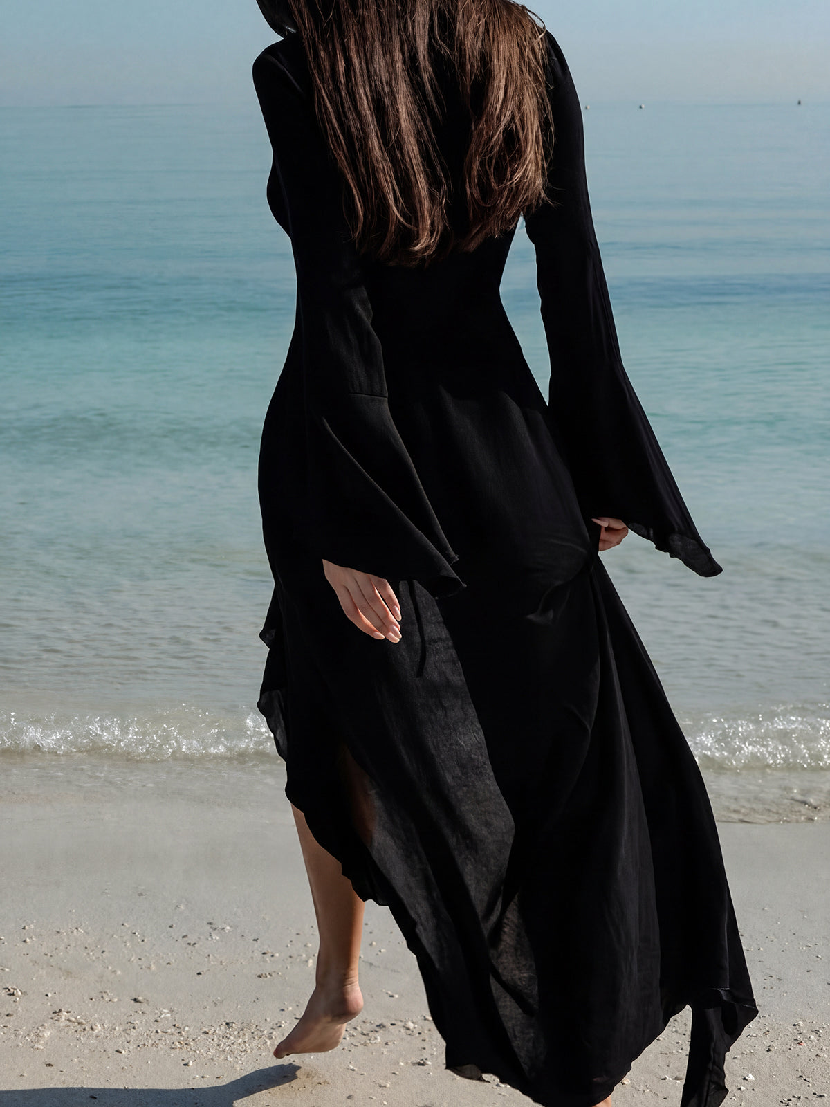 Woman walking away from the camera on a beach, wearing a long-sleeved black ruffled maxi dress.