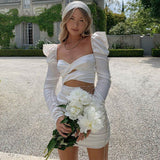 Woman in a white satin dress with long sleeves and a sweetheart neckline, holding a bouquet of white flowers.