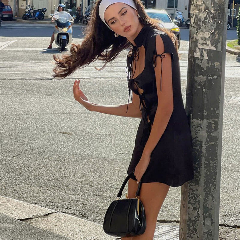 Woman in a black bodycon dress with cut-out details and tie closures, leaning against a pole on a city street.