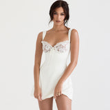 Woman wearing a white mini dress with lace detailing on the bust, looking at the camera with one hand lifting the hem of the dress