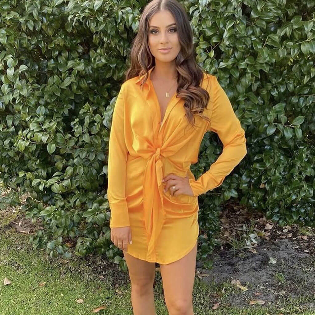 Woman wearing a yellow satin tie-front mini dress standing in front of a green hedge.