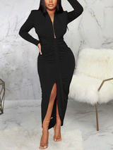 Woman in a black long-sleeved ruched maxi dress with a high slit, standing in a room with marble walls and a furry chair.