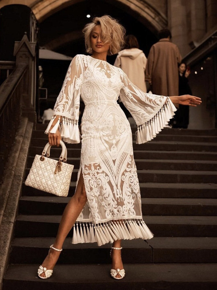 woman wearing a white lace dress with bell sleeves and fringe detail, holding a white handbag and wearing white heels, standing on stairs