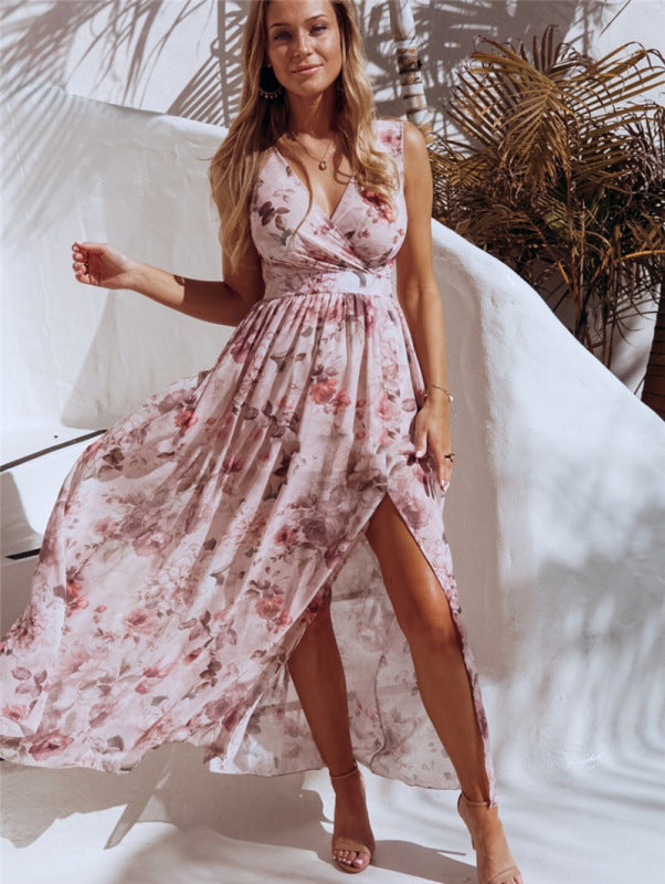 Woman wearing a pink floral print maxi dress with a high slit, posing in an outdoor setting with white curved structures and palm leaves.