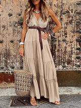 A woman stands in a beige flowing maxi dress, highlighting its sleeveless cut, V-neckline, tiered design, and subtle lace trim, complemented by casual accessories.