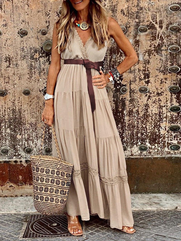A woman stands in a beige flowing maxi dress, highlighting its sleeveless cut, V-neckline, tiered design, and subtle lace trim, complemented by casual accessories.