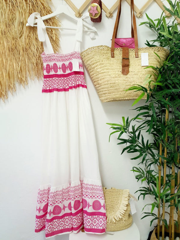 A white and pink embroidered maxi dress hanging on a wall, next to a straw bag and plants.