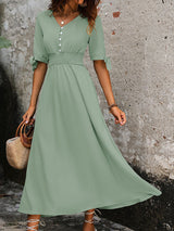 Woman wearing a sage green A-line maxi dress with a smocked waist and tie sleeves, standing in front of a textured wall.