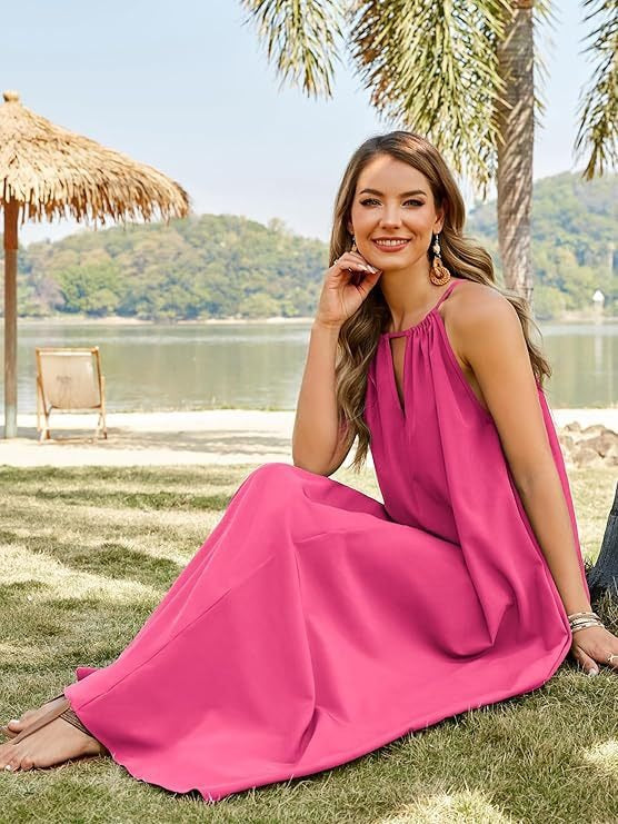 Woman wearing a bright pink sleeveless maxi dress with a halter neckline and keyhole cutout, sitting on the grass by a body of water with palm trees in the background.