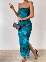 Woman posing with a black clutch, wearing a strapless blue and green abstract floral print maxi dress with a high slit and gold heeled sandals.