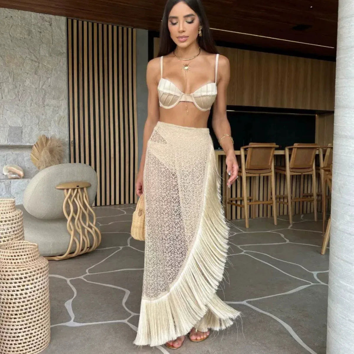 Woman wearing a beige bikini set with a sheer lace maxi skirt with fringe detailing, standing indoors with modern decor.