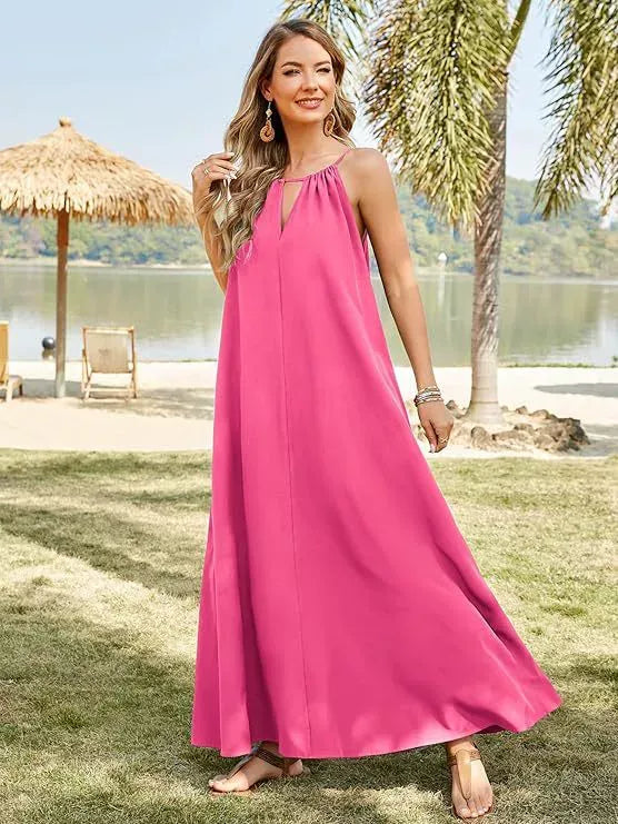 Woman wearing a bright pink sleeveless maxi dress with a halter neckline and keyhole cutout, standing on grass by a body of water with palm trees and a thatched umbrella in the background.