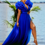 Woman wearing a flowing royal blue maxi dress with ruffled shoulders and a high slit, standing on a beach with palm trees and water in the background.