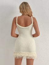 Back view of a woman wearing a white knit mini dress with fringe hem and braided straps, standing against a white wall.
