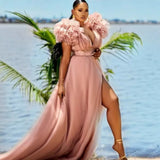 Woman wearing a flowing pink maxi dress with ruffled shoulders and a high slit, standing on a beach with palm trees and water in the background.