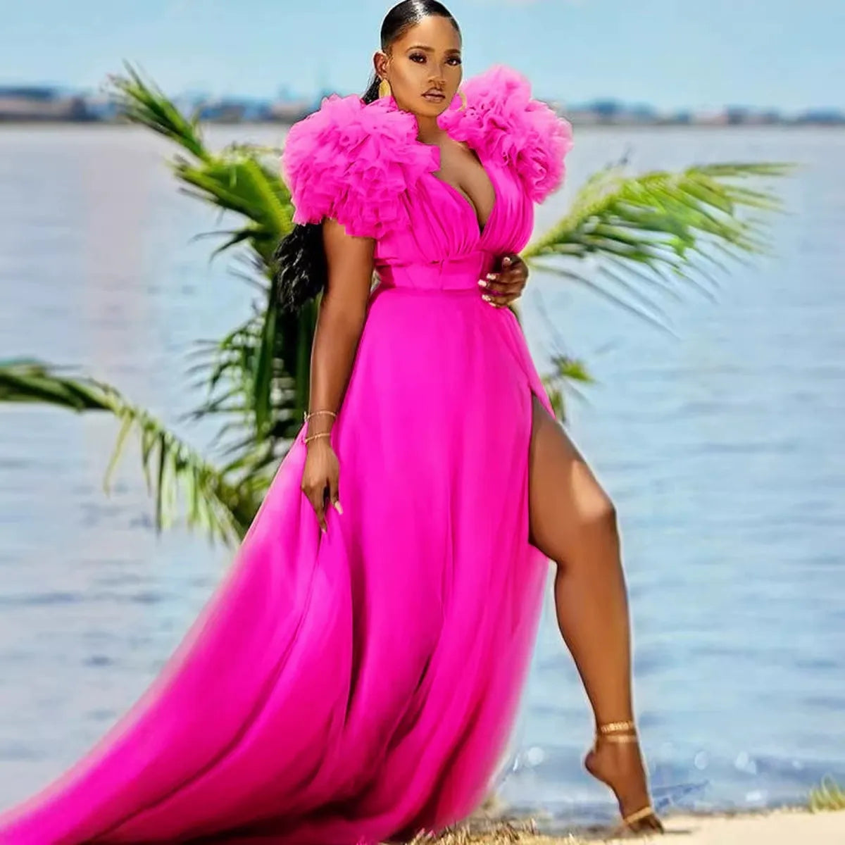 Woman wearing a flowing hot pink maxi dress with ruffled shoulders and a high slit, standing on a beach with palm trees and water in the background.
