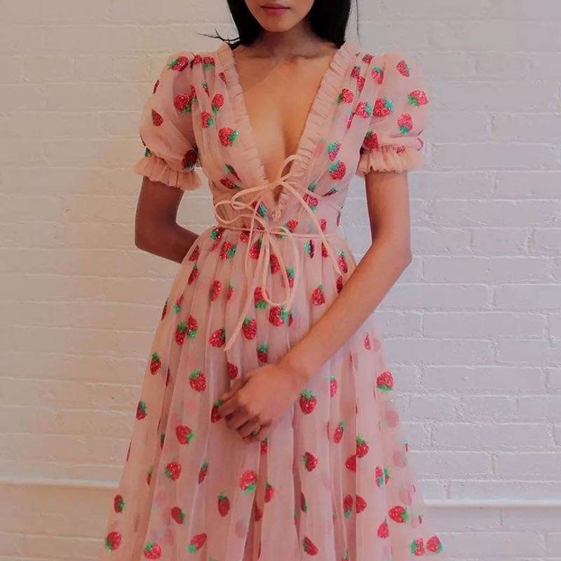 Woman wearing a light pink midi dress with a strawberry print and puff sleeves against a white brick wall.