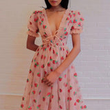 Woman wearing a light pink midi dress with a strawberry print and puff sleeves against a white brick wall.