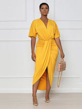 Woman wearing a yellow V-neck, short-sleeve, ruched, draped midi dress with a tulip hem, holding a beige handbag.