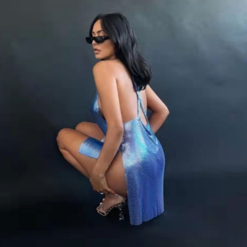 Woman in a blue metallic mesh mini dress with a cowl neckline and high slit, crouching against a dark background.