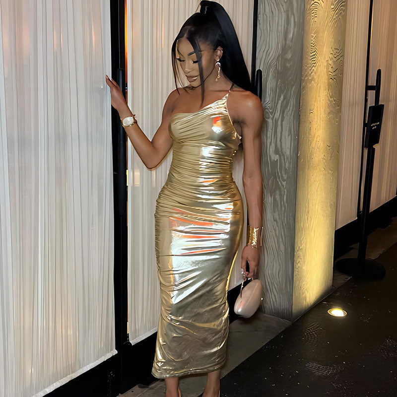 Woman in a gold metallic one-shoulder ruched maxi dress standing near a textured wall.