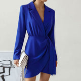 Woman wearing a royal blue blazer dress with a wrap front and tie detail, holding a white clutch.