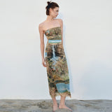 Model wearing a strapless, full-length dress with a landscape print, standing on sand against a white wall.