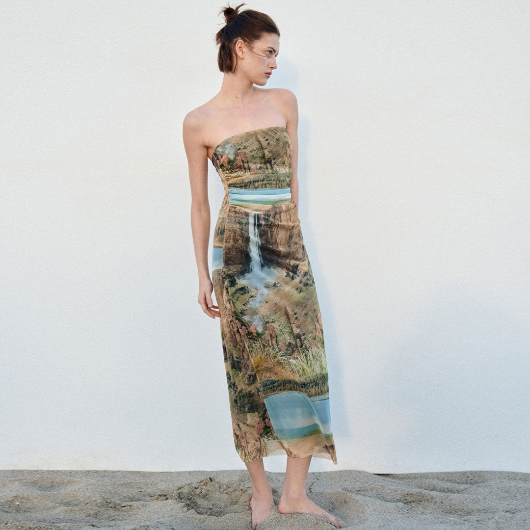 Model wearing a strapless, full-length dress with a landscape print, standing on sand against a white wall.