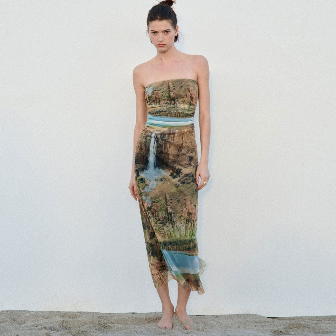 Full-body shot of a model wearing a strapless, full-length dress with a landscape print, facing the camera.