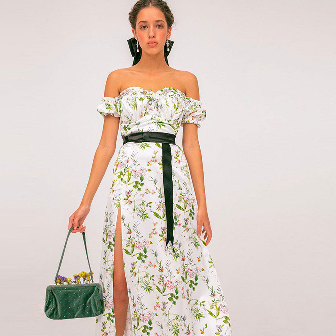 Woman in a white floral off-the-shoulder maxi dress with a high slit and black belt, holding a green handbag.