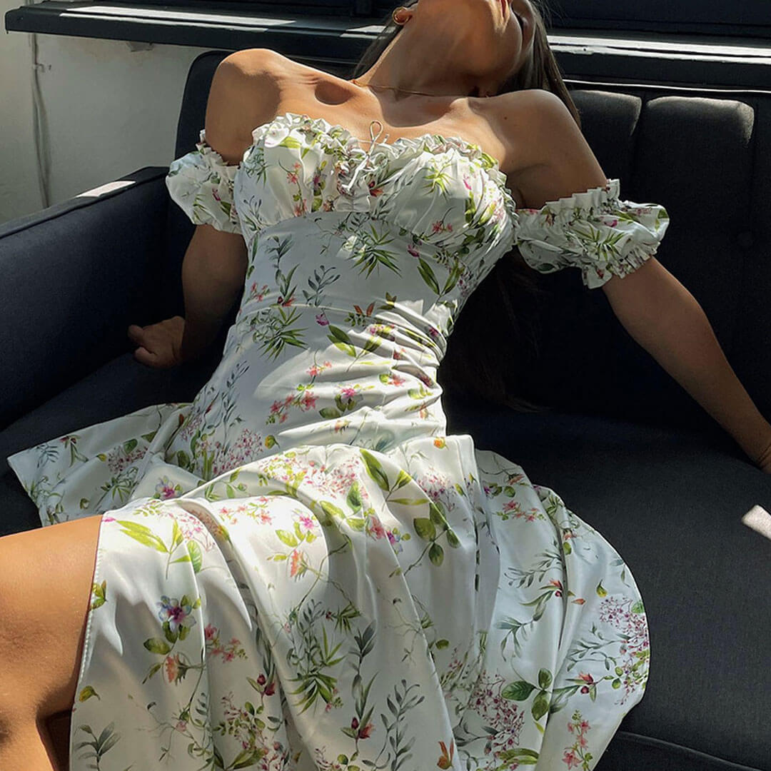 Woman in a white floral off-the-shoulder maxi dress with a high slit, lounging on a sofa.