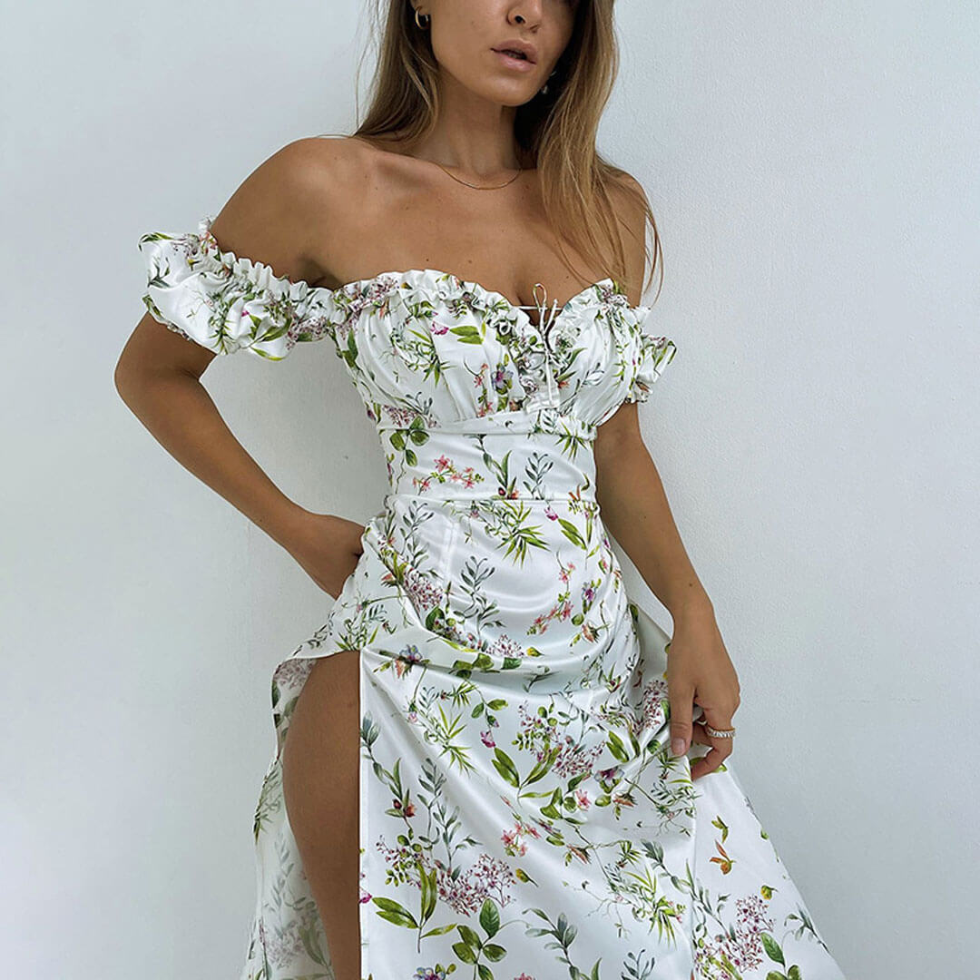 Woman in a white floral off-the-shoulder maxi dress with a high slit, standing against a white wall.