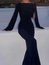 Woman in a black long-sleeved maxi dress with flared sleeves, leaning on a white ledge with a small handbag.