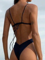 Back view of a woman wearing a navy blue one-piece swimsuit with thin straps and a high-cut leg.