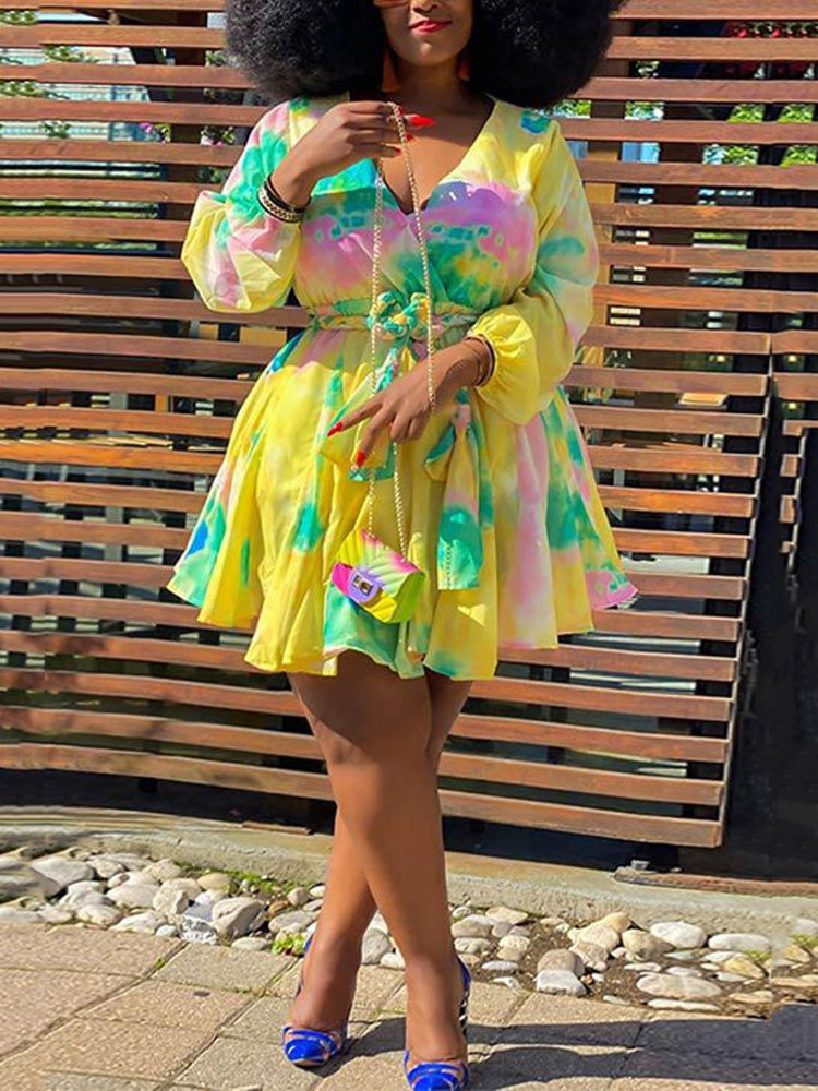 Woman wearing a yellow tie-dye mini dress with a V-neck and full sleeves, accessorized with a small colorful purse and blue heels, standing outdoors.