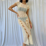 Woman wearing a sheer cream midi dress with short sleeves and tie closures on the side, standing in front of a white curtain.