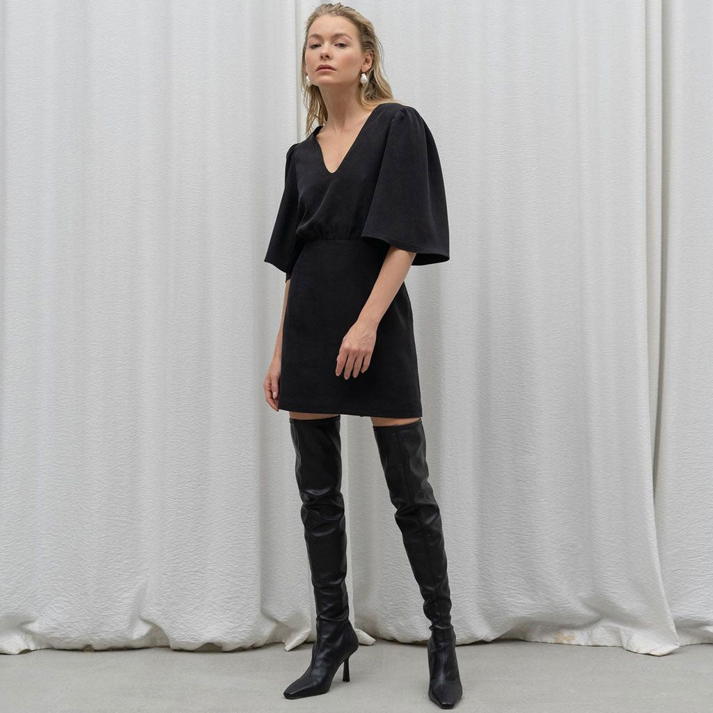 Model wearing a black mini dress with flutter sleeves and black knee-high boots, standing against a white backdrop.