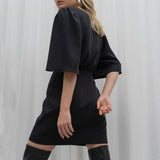 Back view of a model wearing a black mini dress with flutter sleeves and black knee-high boots.