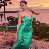 Woman wearing a green satin halter neck maxi dress with cut-out details and a tie-back closure, standing on a hill overlooking the ocean at sunset.