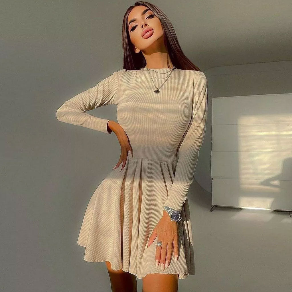 Woman wearing a long-sleeved beige ribbed knit fit and flare dress with a crew neck and cut-out detail, standing with one hand on her hip.