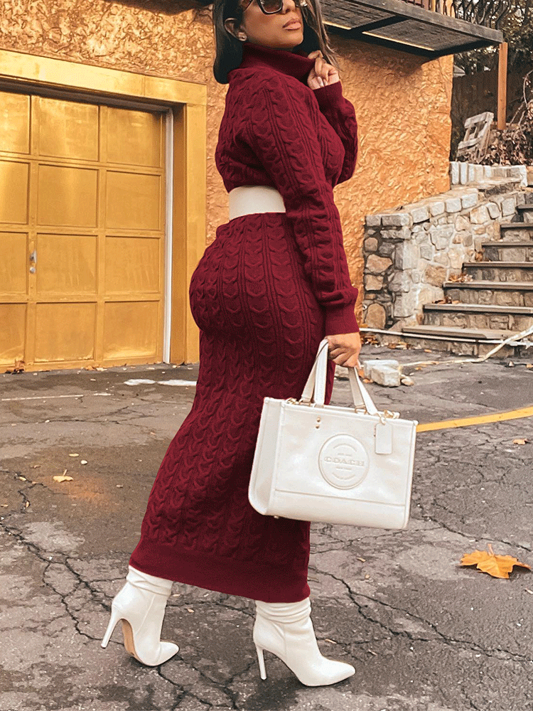 Side view of a woman in a burgundy cable knit turtleneck maxi dress with a white belt and white boots, holding a white handbag.