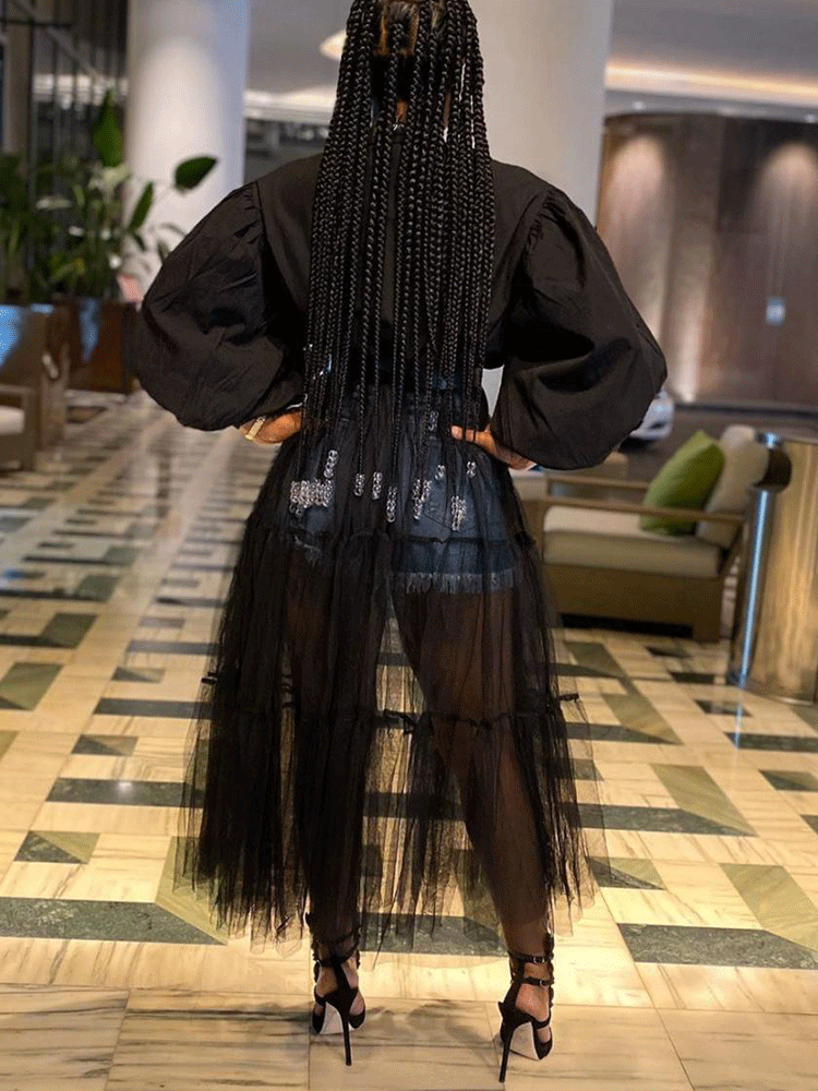 Rear view of a woman wearing a black shirt dress with a sheer black tiered tulle skirt overlay and denim shorts, with braided hair.