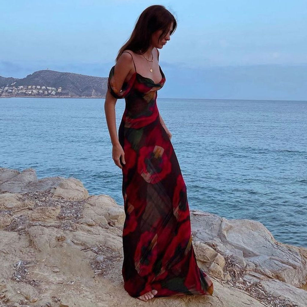 Woman in a long, off-the-shoulder dress with a floral print, standing on rocks near the ocean at dusk.