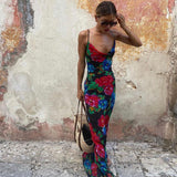 Woman wearing a black floral print maxi slip dress with a cowl neckline and spaghetti straps, holding a woven tote bag.